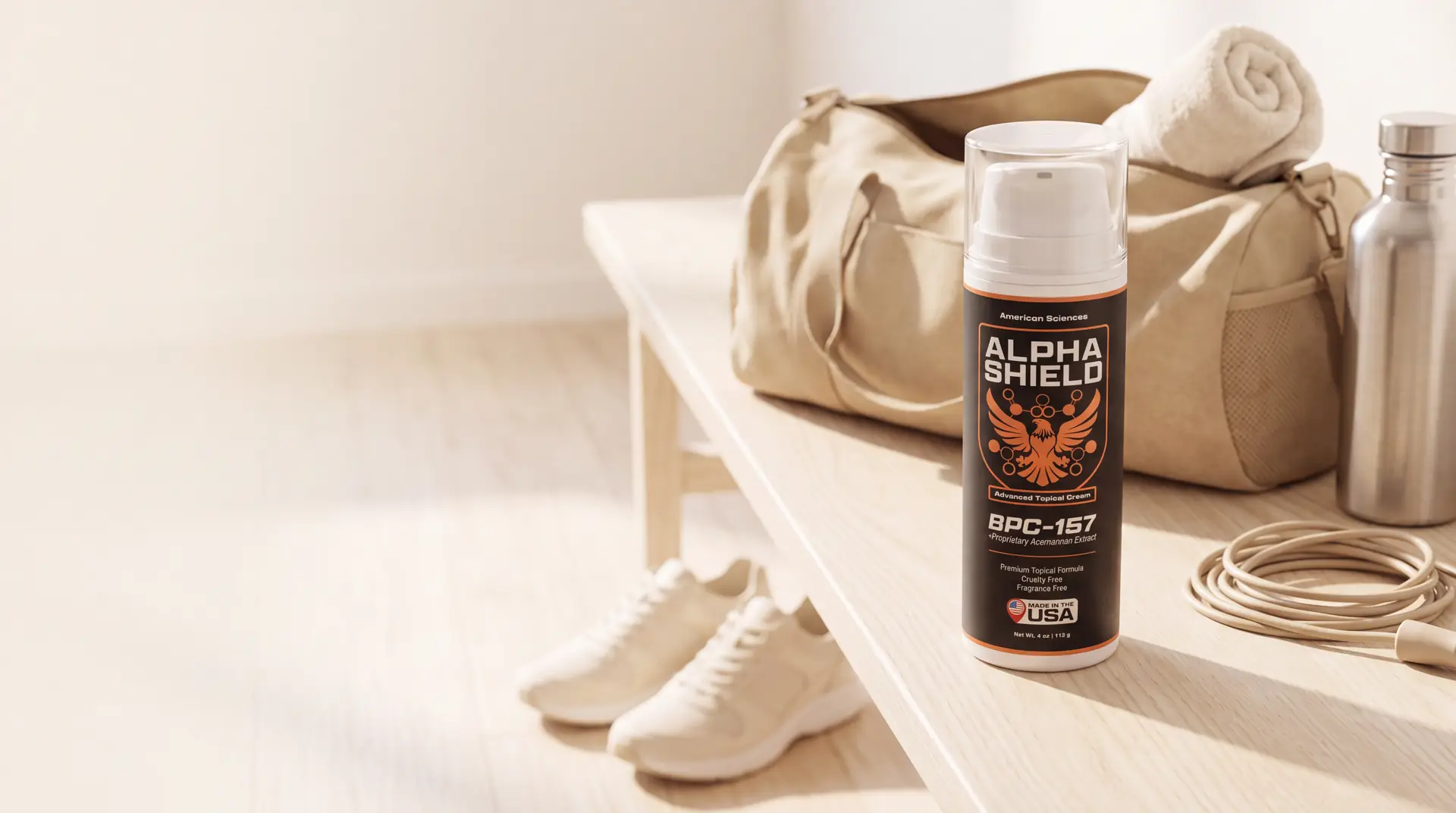 Alpha Shield bottle on a pale wooden bench in a sunlit home gym, beside an open canvas gym bag, a rolled towel and a steel water bottle